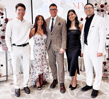 Five well-dressed individuals posing together at an event with floral decorations.