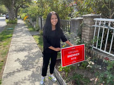 A woman stands smiling next to a political campaign sign in a sunny neighborhood.