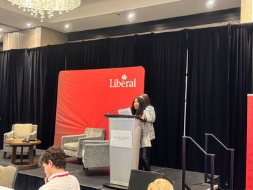 Woman speaking at a Liberal event podium with audience and chairs.