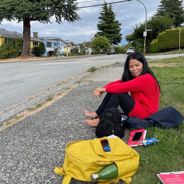 A woman in a red sweater sits barefoot on the sidewalk with bags and books nearby.