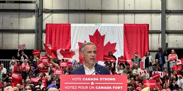 A political rally with a speaker in front of a large Canadian flag and crowd holding signs.