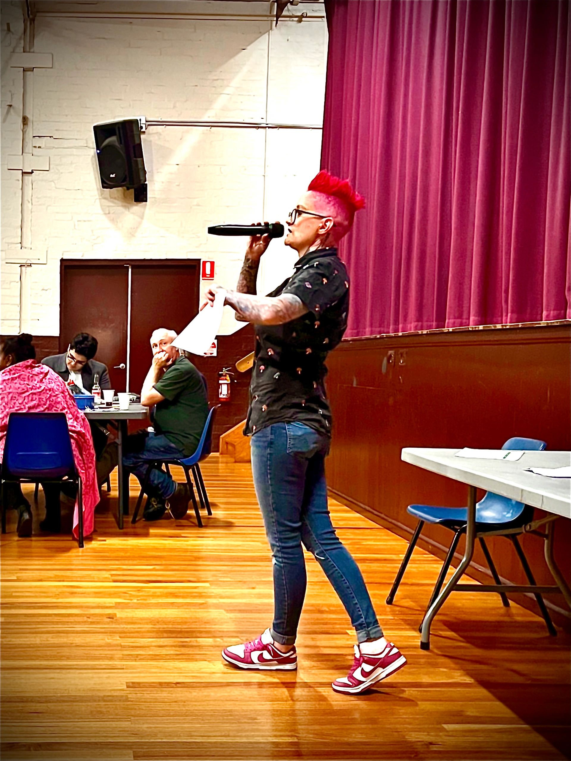 Person with bright red mohawk speaking into a microphone on a wooden floor stage.