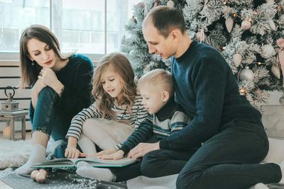Family of four reading a book by a decorated Christmas tree.