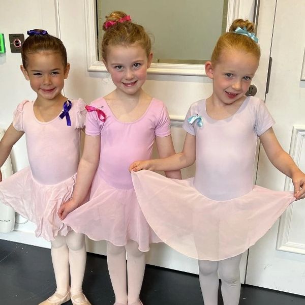 Three young girls in pink ballet outfits posing happily indoors.