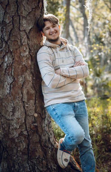 Young man leaning against a tree smiling warmly outdoors.