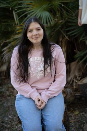 Young woman sitting outdoors in a pink sweatshirt and blue jeans.