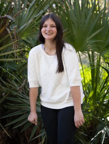 Young woman in white sweater smiling outdoors by green foliage.