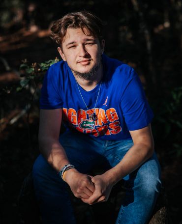 Young man in a blue shirt sitting outdoors with clasped hands.