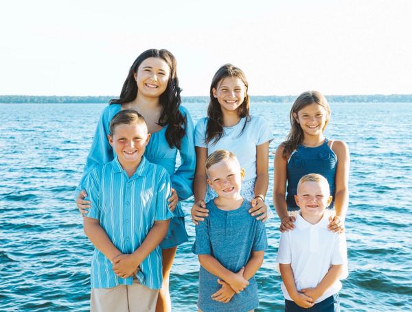 A smiling group of six children posing by the water on a sunny day.