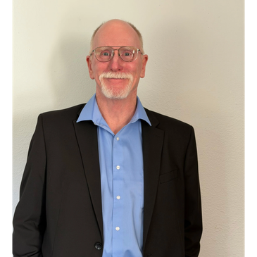 Smiling elderly man in glasses wearing a blue shirt and black blazer.