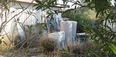 Overgrown backyard with old appliances and scattered debris next to a white shed.