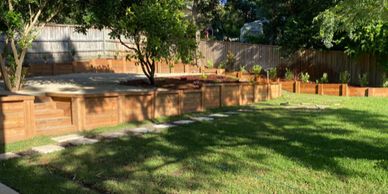 A sunny backyard with a large green lawn and wooden retaining walls.
