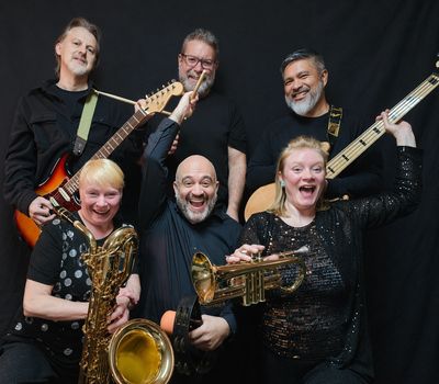 Energetic band with diverse instruments posing joyfully against a black backdrop.