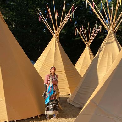 Person in traditional attire stands among several teepees in sunlight.