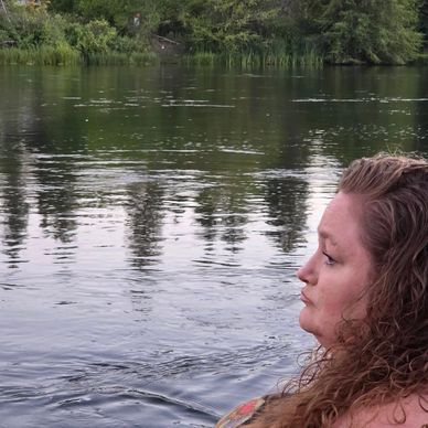 Woman with curly hair gazes thoughtfully at a calm lake surrounded by trees.