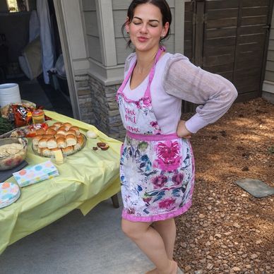 Woman in floral apron posing by a picnic table with food outdoors.