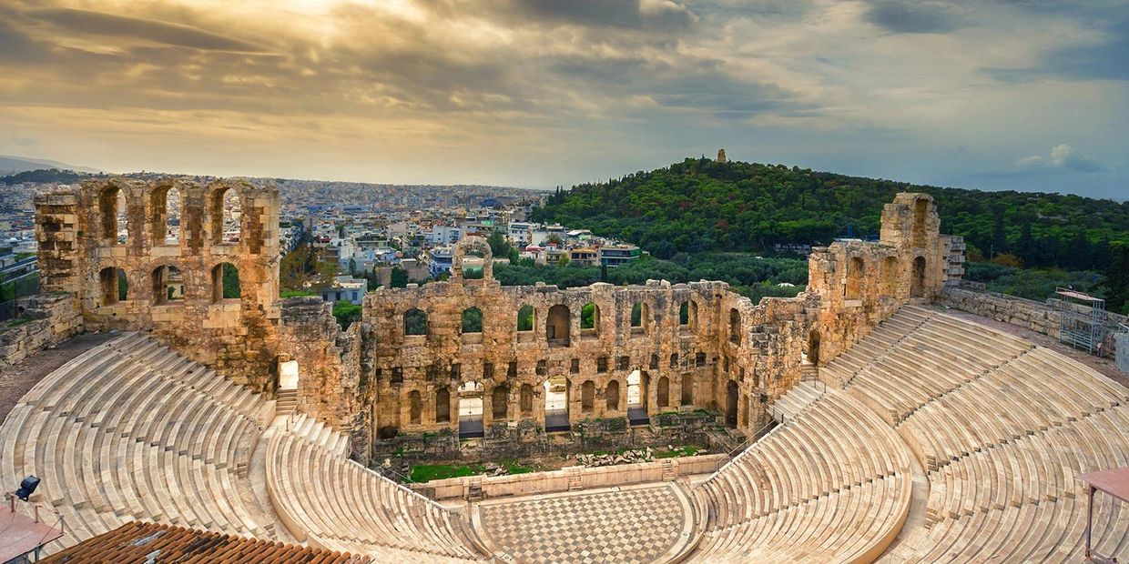 Ancient stone amphitheater with city and forest in background under a cloudy sky.