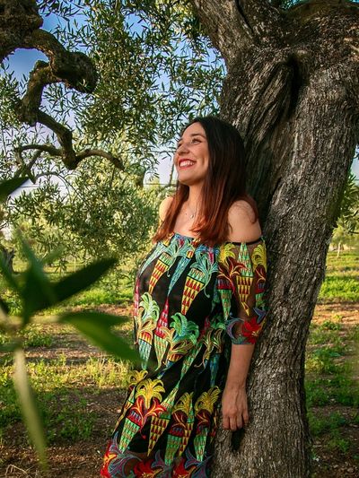 A smiling woman in a colorful dress leans against an old tree in a green field.