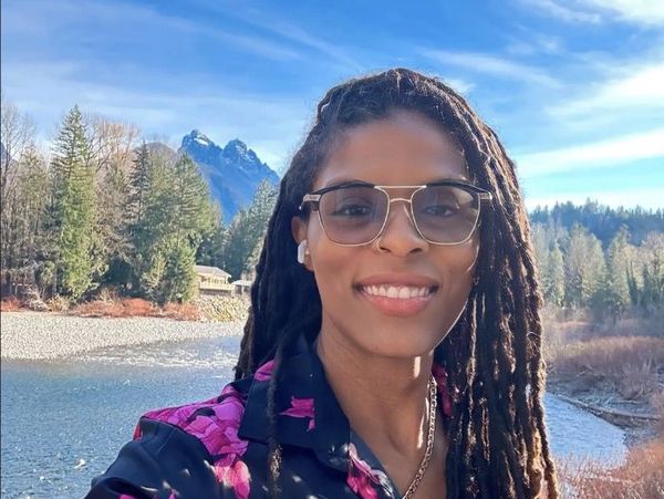 Woman smiling with glasses in a floral shirt by a river and mountains.