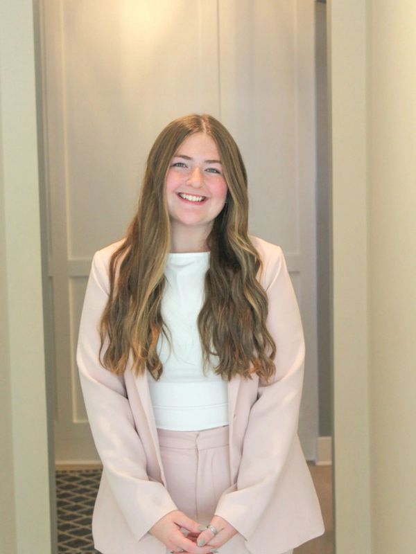 A smiling young woman with long hair wearing a light pink suit and white top.