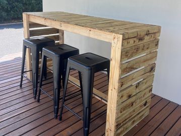 Outdoor wooden bar table with three black metal stools on a wooden deck.