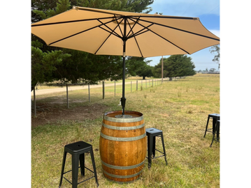 Outdoor setup with a wooden barrel table, beige umbrella, and black stools on grassy field.