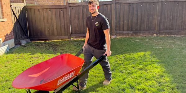 A man smiling and holding a red wheelbarrow in a sunny backyard.