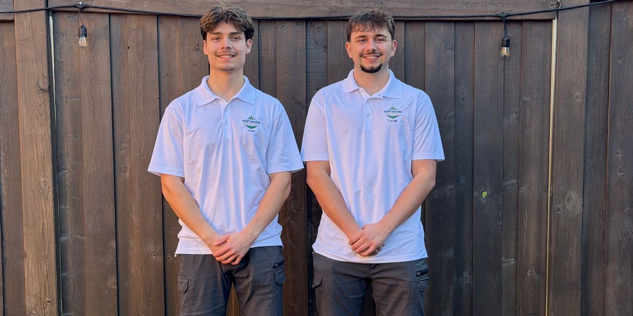 Two young men in white polo shirts standing in front of a wooden fence.