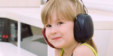 A young child wearing large earmuffs, smiling indoors.