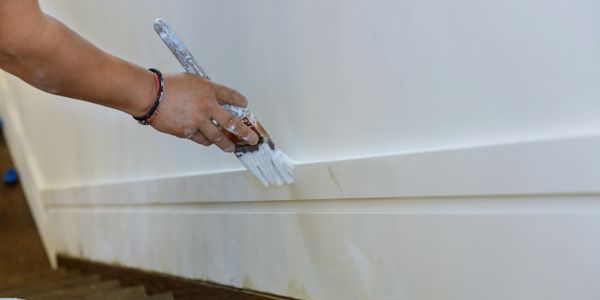 A person painting a white baseboard with a paintbrush.