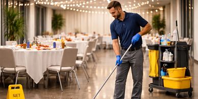 Janitor cleaning debris on the floor in a banquet hall with a broom and cleaning cart.
