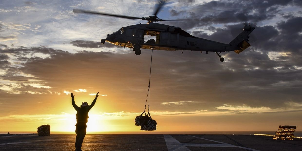 Seahawk helicopter dropping cargo on the deck of an aircraft carrier at sunset.