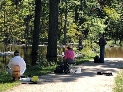 Three people painting outdoors by a peaceful wooded pond on a sunny day.