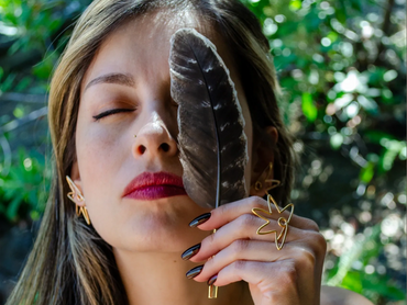 Woman holding a feather over one eye with eyes closed in a natural setting.