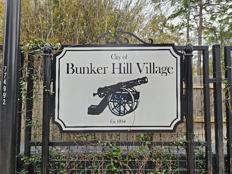 City of Bunker Hill Village sign with a black canon. Black fence with trees in the background.