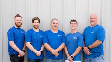 Five men in matching blue company shirts pose against a metal wall.