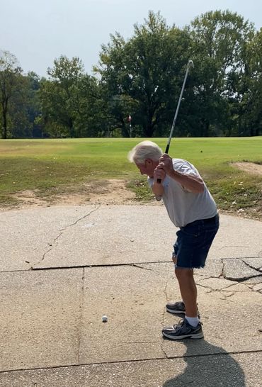 Older man preparing to hit a golf ball on a cracked concrete surface near a green.