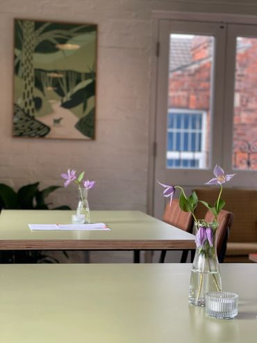 Purple flowers in glass vases on tables in a cozy room with a window and artwork.