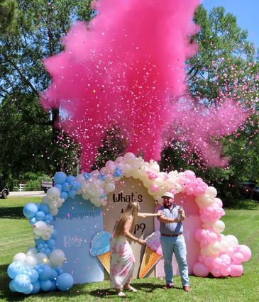 A gender reveal party with pink smoke and balloons in a park.
