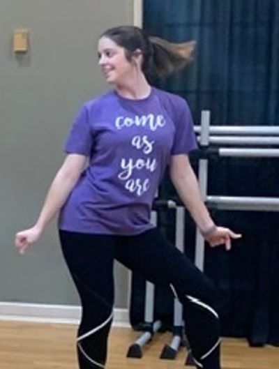 Woman dancing indoors wearing a purple shirt saying 'come as you are'.