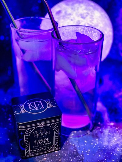 A tin of Sunrise Sencha tea in front of two clear glasses and a violet moon.