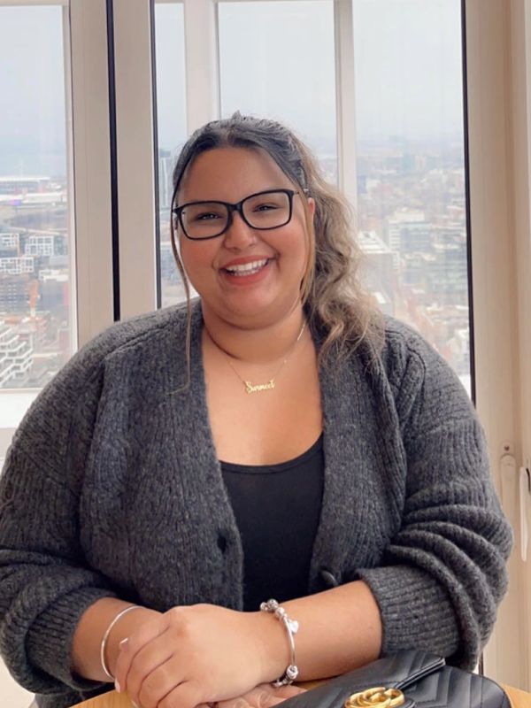 Smiling woman in glasses and cozy grey sweater sitting indoors by a window.