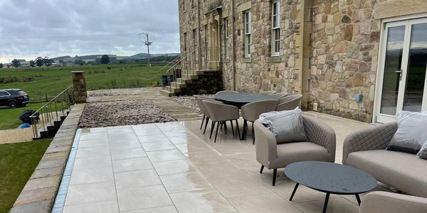 Large light patio with seating, dining, and drainage next to stone house in North East UK garden.