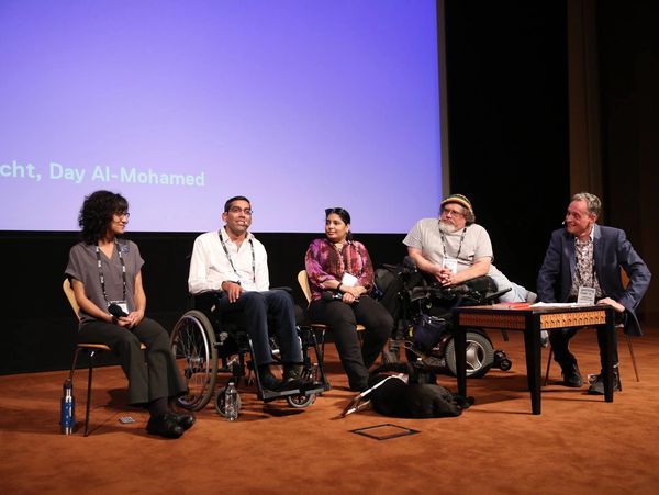 Five panelists sitting in chairs and wheelchairs on a stage in front of a screen