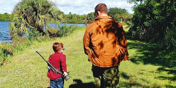 A young boy in a red plaid shirt and jeans carries a rifle over his shoulder while walking with Dad