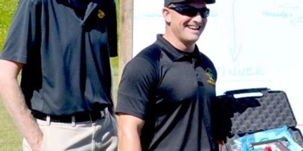 Two men wearing black shirts and sunglasses stand outdoors. The man on the right laughs