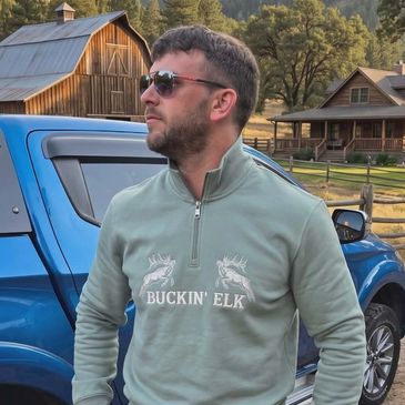 Man wearing sunglasses and a green pullover stands by a blue truck in a rustic setting.