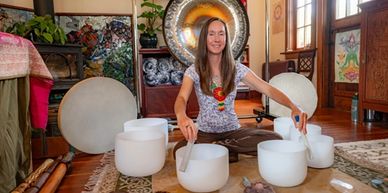 A woman plays crystal singing bowls in a cozy room filled with plants and artwork.