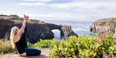 Woman practicing yoga by the ocean with a natural rock arch in the background.