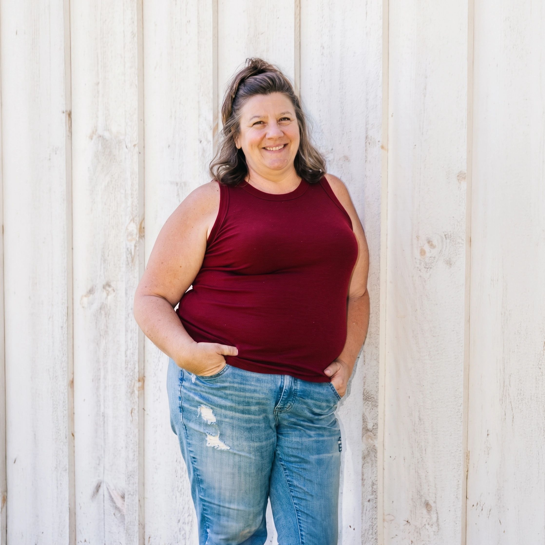Smiling woman in a red sleeveless top and jeans standing against a white wooden wall.